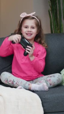 Küçük kızın evdeki oturma odasında video çalarkenki dikey videosu. Heyecanlı oyuncu kız elinde joystick ile kablosuz kumanda kullanarak konsol oyunu oynuyor. Eğleniyor, mutlu ifadelerin tadını çıkarıyor..