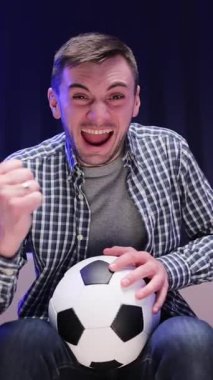Topu elinde tutan adamın dikey format videosu spor kanalını izliyor. Yakışıklı beyaz adam endişeleniyor ve futbol maçı için neşeleniyor. Gece geç saatte kanepede oturuyor..