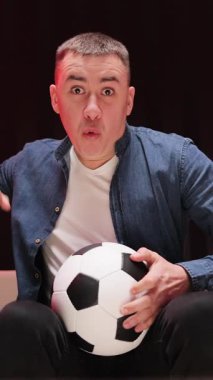 Gece geç saatlerde kanepede oturan yakışıklı, endişeli ve futbol maçı için neşelenen beyaz adamın dikey format videosu. Elinde top olan adama yaklaş ve spor kanalını izle..