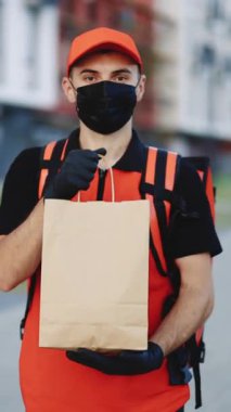 Kırmızı üniformalı, şapkalı ve tıbbi maskeli beyaz adamın dikey format videosu servis yaparken kameraya yemek paketi ile gülümsüyor. Çevre marketi paketli erkek kurye..