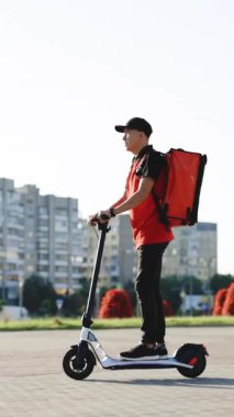 Asyalı Kurye 'nin kırmızı termal sırt çantalı kurye yemek teslimatının dikey videosu elektrikli scooterla sokakta müşteriye sipariş verirken görülüyor. Kırmızı üniformalı teslimatçı işçi..