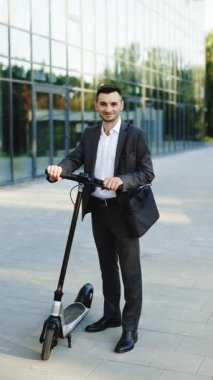 Açık havada duran zarif takım elbiseli genç işadamının dikey format videosu. Neşeli bir şekilde gülümsüyor ve başparmağını gösteriyor. İş binasının önünde elektrikli scooterıyla yakışıklı bir iş adamı..