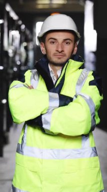 Ağır endüstri işçisinin Zoom portresinin dikey videosu. İnşaat alanında çalışırken kameraya güvenerek gülümsüyor. Yüksek voltajlı elektrik santrali. Elektrikçi, elektrik işçisi.