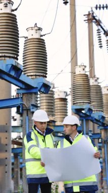 İki mühendisin dikey format videosu, yüksek voltajlı bir elektrik hattının arka planına karşı açık havada kağıt üzerine bir çizimi tartışıyor. Özel kıyafetli mühendisler olay yerinde çalışıyor..