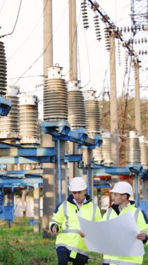 Elektrik hattı işçilerinin günbatımı arka planında bir inşaat planıyla çalıştıkları dikey video. İki mühendis, yüksek voltajlı elektrik hattının arka planına karşı açık havada kağıt üzerine çizim yapmayı tartışıyor..