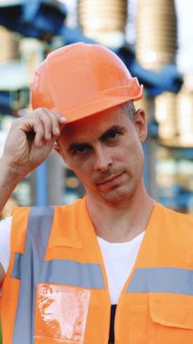 Dikey Format Video: Genç Profesyonel Ağır Sanayi Mühendisi İşçi Çelik Yelek Giyiyor, Sıkı Şapka Giyiyor. Arkaplanda Odaklanmamış Büyük Güç İstasyonunda