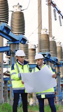 Beyaz kasklı ve yeşil yelekli iki mühendisin dikey format videosu yüksek voltajlı bir istasyonun önünde duruyor..