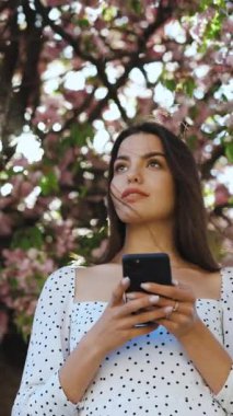 Sakura ağaçlarının arka planında akıllı telefon kullanan güzel bir kadının portresinin dikey format videosu. Sosyal ağları kullanarak internette gezinen güzel bir kız..
