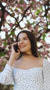 Beyaz bir yaz elbisesi giymiş, arka planda sakura ağaçlarının üzerinde telefonda konuşan manken kızın dikey videosu. Uzun saçlı bir kız akıllı telefondan konuşur ve dışarıda gülümser..