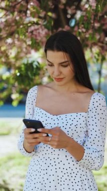 Sakura ağaçlarının arka planında akıllı telefon kullanan güzel bir kadının dikey format videosu. Sosyal ağları kullanarak internette gezinen güzel bir kız..