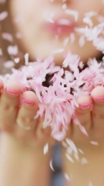 Dikey Portre 'nin muhteşem kızı elinde pembe çiçek yaprakları tutuyor. Dalgalanan yapraklar, rüzgarlı hava. Elinde çiçek açan pembe sakura çiçeklerini üfleyen kız kameraya bakıyor ve gülümsüyor..