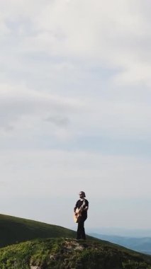 Günbatımında günbatımında müzik aleti çalan müzisyenin dikey videosu. Dağlarda akustik enstrüman çalan genç müzisyenlerin hava görüntüsü. Doğada akustik gitar çalan adam..