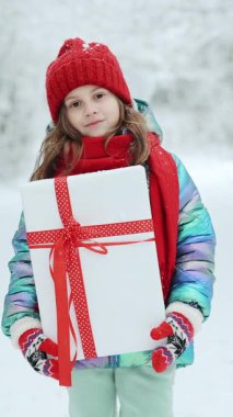 Küçük bir kızın Noel kutusuyla birlikte Noel arifesinde dışarıda geçirdiği dikey video. Küçük mutlu beyaz kız gülümsemesi ve Noel günü hediye paketi taşıyor. Çocuk hediye kutusu ve sürpriz yüz tutuyor..