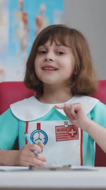 Evde kameraya gülümseyen küçük bir kızın dikey videosu, tıbbi giysiler giyen sevimli bir çocuk web kamerasıyla konuşuyor çevrimiçi video görüşmesi yapıyor ya da video bloğu kaydediyor, güzel yüzlü bir çocuk..