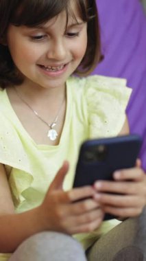 Tatlı kızın telefonunun olduğu dikey format videosu, akıllı telefon kullanan çocuk, internette gezinen çocuk, telefonda video iletişimi kullanıyor..