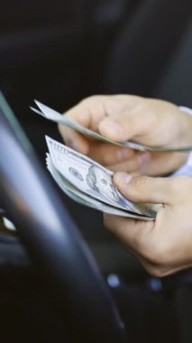Adamın arabada oturmuş dolarları saydığı dikey video. Erkek eli para sayar. Kazanç ya da rüşvet kavramı, sigorta ya da kredi, yatırım. Lüks arabada para sayan bir adam..
