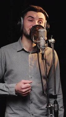 Profesyonel beyaz erkek şarkıcının dikey format videosu. Kulaklık takıyor ve mikrofon kullanarak karanlık iç mekan ile müzik stüdyosunda şarkı söylüyor..