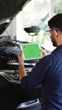 Vertical Video of Car Service Mechanic Uses Tablet Computer with Green Screen Mock Up Chroma Key that is Pointed at an Enginer Bay. Specialist Inspecting the Vehicle in Order to Find Broken Components