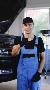 Vertical Video of car mechanic in car workshop shows thumbs up. Positive auto service worker smiling to camera and showing thumb up gesture, approving car repair workshop. Workday. Repair concept.