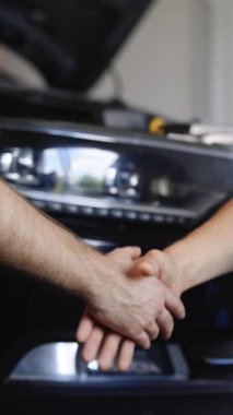 Tamircide el sıkışan tamirci ve müşterinin dikey videosu. Tanımlanamayan iki adam otomobil tamircisi ve müşterisi araçların arka planında el sıkışıyor. Araba işi, tamir hizmetleri.