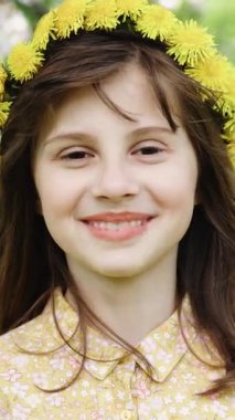 Yazın başında çelenk olan mutlu gülümseyen bir kızın dikey videosu. Kız yüzü parkta. Bir çocuğun mutlu yüzü. Çocuk kameraya gülümser. Yüzü yakın plan. Parktaki mutlu çocuk. Güzel yüz.