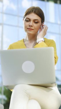 Diz üstü bilgisayar kullanarak bankta otururken en sevdiği müziği kablosuz kulaklıkla dinleyen güzel beyaz kadının dikey videosu. Mutlu Avrupalı kız Mola ve yumuşak dansın tadını çıkarıyor.