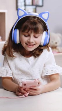 Akıllı telefondaki komik kurgu uygulamasını kullanan, sosyal ağdaki havalı video veya müzik içeriğinden hoşlanan, çevrimiçi oyunlar oynayan, uzaktan iletişim kuran, kulaklıklı şirin küçük bir kızın dikey videosu.