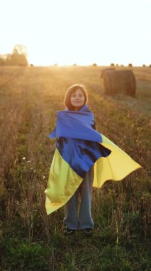 Gökyüzünde mavi-sarı bayrak taşıyan Ukraynalı bir kızın dikey format videosu. Ukraynalı bir kızın portresi Ukrayna bayrağıyla tarlanın ortasında gün batımına karşı duruyor.