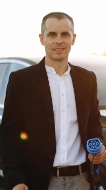 Dikey Format Video: Güvenilir genç profesyonel iş adamı lider kazanan çalışan kameraya bakıyor. Açık hava elektrikli arabanın yanındaki şarj istasyonunda el işareti hareketi gösteriyor..