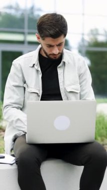 Modern ofis yakınlarındaki dizüstü bilgisayarda çalışan sakallı iş adamının dikey videosu. İş yerinde dizüstü bilgisayarda daktilo kullanan profesyonel bir erkek. Dışarıdaki dizüstü bilgisayara bakan pozitif iş adamı.