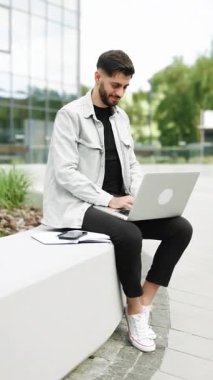 Ofis dışında dizüstü bilgisayara bakan bir adamın dikey görüntüsü. Modern ofisin yanındaki bankta oturan gülümseyen adam. İş adamı uzak işyerindeki laptop kamerasına gülümsüyor