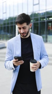 Modern şehir bölgesinde cep telefonu kullanan kendine güvenen bir Ermeni 'nin dikey videosu. Pozitif iş adamı açık havada gülümseyen finans haberlerine bakıyor. Başarılı bir yetişkin adam..