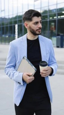 Tablet ve kahve bardağıyla iş binaları arasında yürüyen ve neşeyle gülümseyen başarılı beyaz sakallı genç adamın dikey videosu. Çekici bir iş adamı. Kariyer insanları.