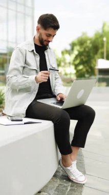 Erkek alıcının modern ofis yakınlarındaki bir internet sitesinden ödeme yaptığı dikey format videosu. Bilgisayarlı e-bankacılık hizmeti kullanan müşteri güvenli online ödeme yaparak kredi kartını elinde tutuyor.