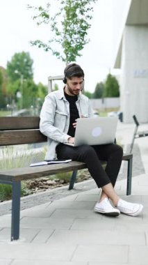 İş adamının kulaklık taktığı, bilgisayardan iş görüşmesi yaptığı ve web kamerasıyla iletişim kurduğu dikey bir video. Erkek Yönetici konuşuyor, bilgisayardan sohbet ediyor ofisin yanındaki bankta oturuyor.
