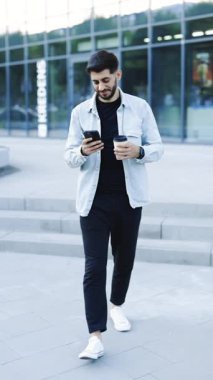 Güzel saç stili ve sakallı, akıllı telefonunu kullanan ve kahve yazan mesajlar yazan çekici sakallı yakışıklı adamın dikey videosu. Çok şık ve muhteşem görünüyorsun..