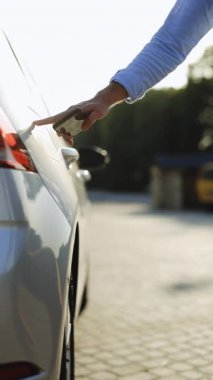 Elektrikli arabayı şarj eden adamın dikey videosu. Akıllı telefondaki uygulamayı kullanarak elektrik kablosuna bağlanan bir erkek. İş adamı gün batımında şarj istasyonunda elektrikli araba şarj ediyor.