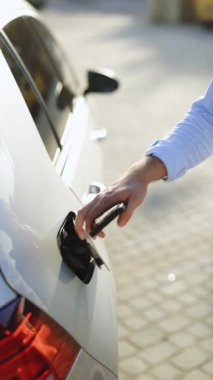 Akıllı telefondaki uygulamayı kullanarak elektrik kablosuna bağlanan bir erkeğin dikey videosu. Elektrikli arabayı şarj istasyonundan çıkaran adam. İş adamı gün batımında şarj istasyonunda elektrikli araba şarj ediyor.