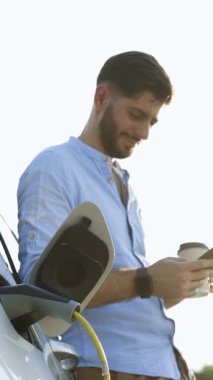 Dikey Format Video: Mavi tişörtlü muhteşem sakallı beyaz adam elektrik yüklü bir arabanın yanında duruyor ve akıllı telefondan zaman ayarları yapıyor..