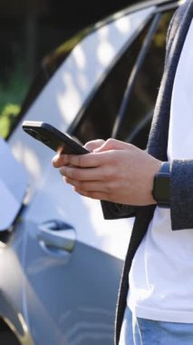 Plug 'ın şarj olan bir arabayı şarj ettiği dikey format video. Beyaz adam akıllı telefon kullanıyor ve güç kaynağı bekliyor. Arabanın aküsünü şarj etmek için elektrikli araçlara bağlanıyor..