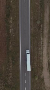 Yarı römorklu, otoban nakliye kamyonunun hava görüntüsünün dikey format videosu. Ağır bir kamyon yeşil tarlalar boyunca asfalt bir yolda gidiyor. Kargo teslimatı, ulaşım.