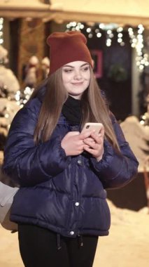 Gece Kasabası 'nın Sokakları' nda yürürken cep telefonu kullanan çekici kadının dikey videosu. Cep telefonundan mesaj atan genç kız. iPhone 'unda APP kullanan şık görünümlü bir kadın. Sosyal Ağ