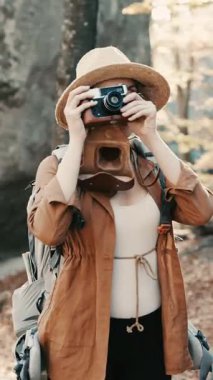 Aktif, beyaz, sırt çantalı bir kadının orman kayalıklarında eski bir kamerayla fotoğraf çektiği dikey format videosu..