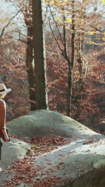 Tepenin tepesinde dağları ve tepeleri olan şapkalı bir kadın gezginin dikey videosu. Sırt çantalı, keçeli şapkalı ve ceketli kız güneşli bir günde etrafa bakıyor..