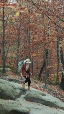 Sırt çantalı çekici kadın turistin dikey format videosu. Boynunda bir kamerayla gezen güzel bir kız, sonbahar ormanlarında kayaları geçiyor.