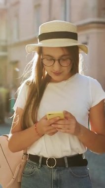 Genç kızın sokakta yürüdüğü ve cep telefonuyla SMS attığı dikey video. iPhone 'unda APP kullanan şık görünümlü çekici bir kadın. Sosyal ağlar. Çevrimiçi olmak. Sanal İletişim