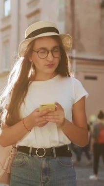Kızın telefonunu kullanırken çekilmiş dikey format videosu. Genç kız, şehirde bir turist dolaşıyor. Sosyal ağlar, çevrimiçi olmak, internette gezinmek. Mesaj atıyorum. Kadın portresi