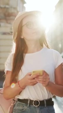 Şehir caddesinde yürüyen ve akıllı telefonundan doğru yöne bakan güzel bir genç kadının dikey format videosu. Mobil Cihazında Çekici Kız Sohbeti. Hasır Şapka Giymek.