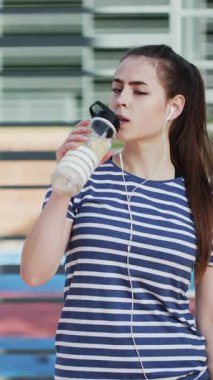 Spor yapan kadının dikey videosu, spor malzemelerinin arka planında Shaker 'dan su içiyor. Spor ve sağlıklı yaşam tarzı. Spor sahasındaki bir parkta spor yapan çekici bir kadın..