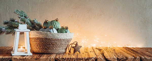 stock image Christmas tree branch and baubles in wicker basket with candle lantern and Christmas lights on wooden table.